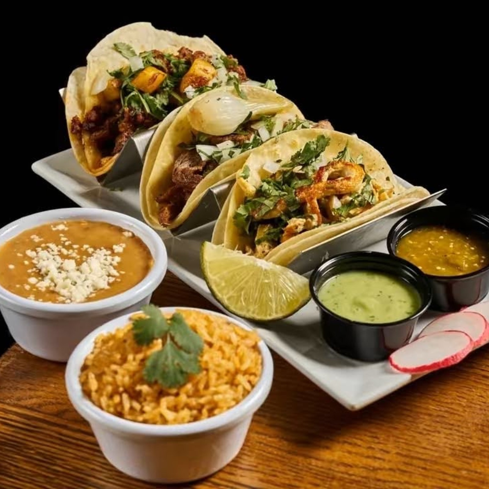 Three loaded tacos with cilantro, onions, lime, rice, beans, and assorted dipping sauces arranged on a restaurant table.