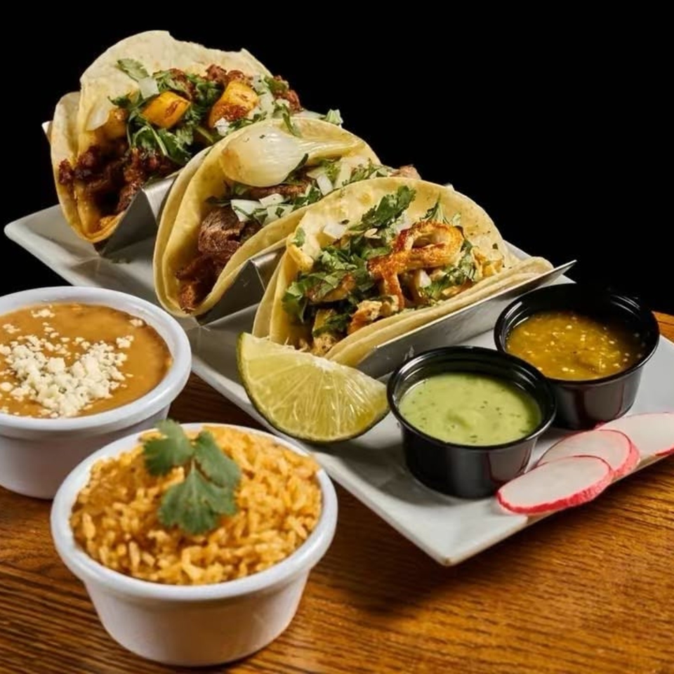 Three loaded tacos with cilantro and onions, Mexican rice, refried beans, lime, radishes, and dipping sauces on a table.