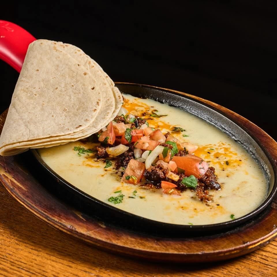 Grilled meat topped with tomatoes, onions, and herbs, served with folded tortillas on a black oval plate.