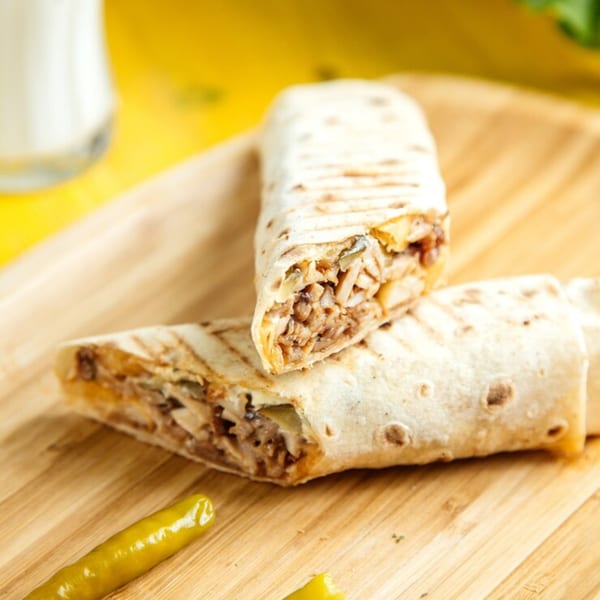 Two grilled burritos filled with meat and vegetables, served on a wooden board with green peppers.