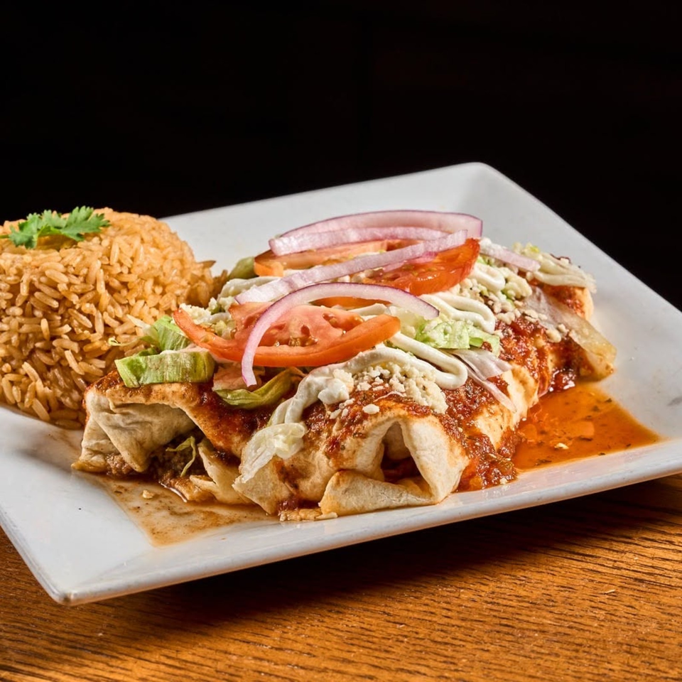Mexican-style enchiladas topped with lettuce, tomatoes, onions, crema, and cheese, served with seasoned rice on a white plate.