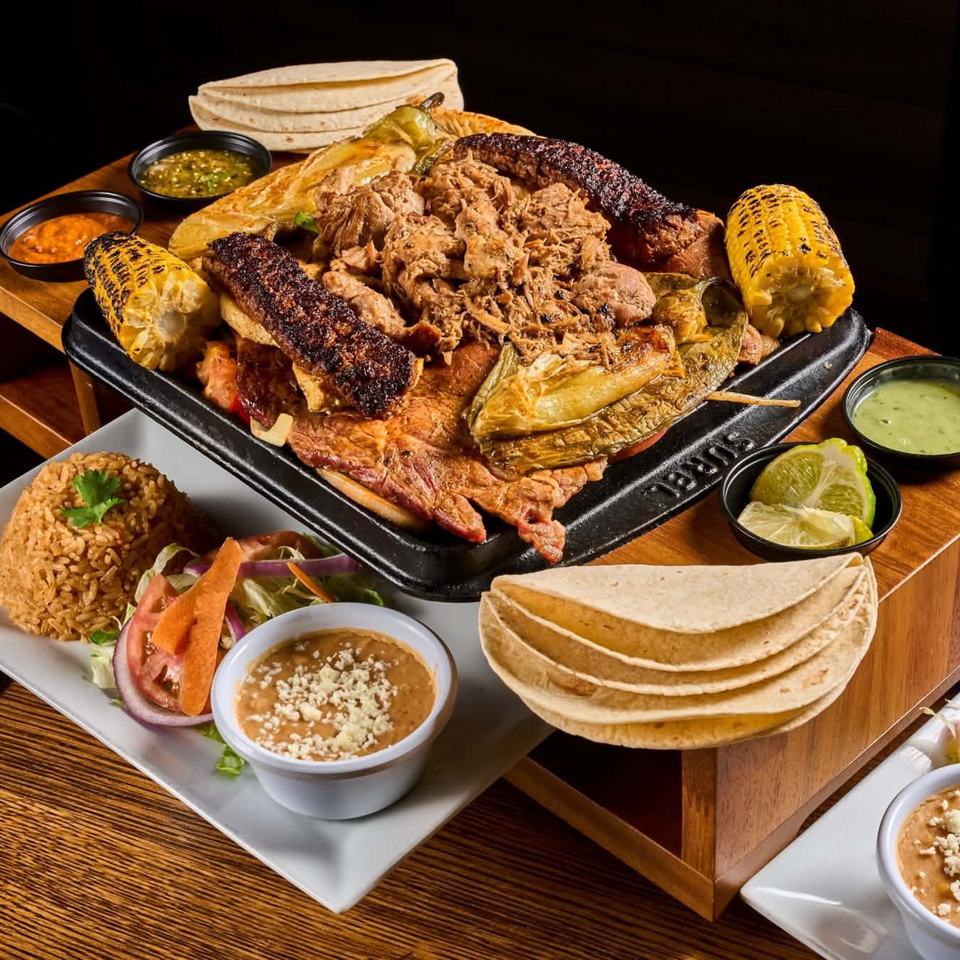 Loaded Mexican parrillada platter with grilled meats, corn, vegetables, tortillas, rice, beans, sauces, and lime wedges.