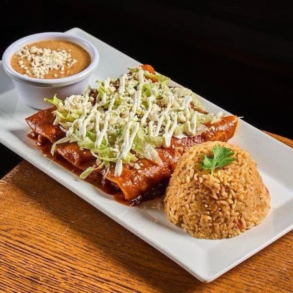 Plate of enchiladas topped with lettuce, cheese, and crema, served with Spanish rice and a side of bean dip.