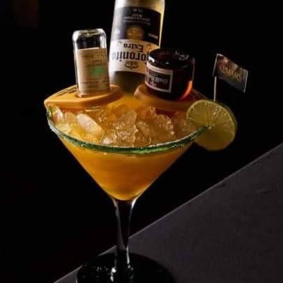 Golden cocktail in a coupe glass garnished with lime, surrounded by liquor bottles and a small flag on a dark bar.
