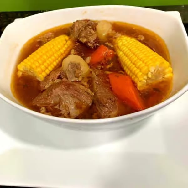 Hearty beef stew with corn on the cob and red bell peppers in a white bowl.