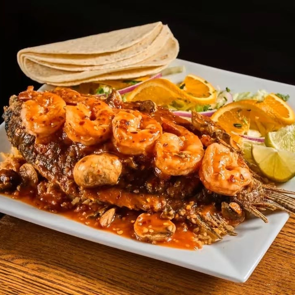 Grilled shrimp and steak skewers in spicy sauce served with tortillas, orange slices, onions, and lime on a white plate.