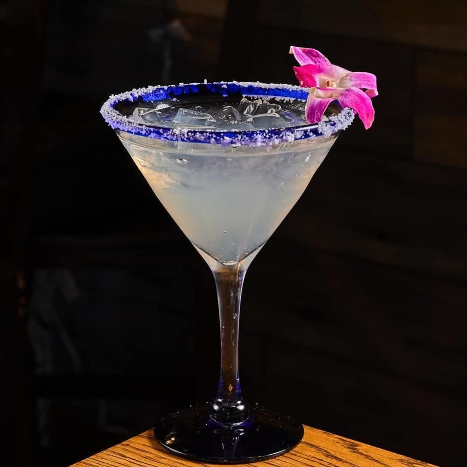 Clear cocktail with ice in a blue sugar-rimmed martini glass, garnished with a bright pink flower against a dark background.